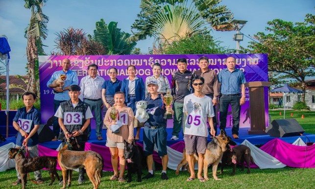 Dog show winners and their dogs standing on stage outdoors with trophies, surrounded by event organizers and judges; several dogs of different breeds are present in front of the group; purple event banner in the background reads International Dog Show Champaign Laos 2023; the atmosphere is celebratory and joyful under a sunny sky with palm trees and greenery around